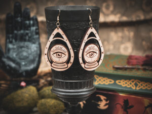 A pair of wooden teardrop-shaped earrings depicting a crystal ball and eye, hanging from a dark metal chalice in a dark academia setting.