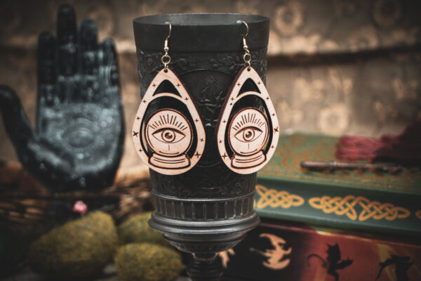 A pair of wooden teardrop-shaped earrings depicting a crystal ball and eye, hanging from a dark metal chalice in a dark academia setting.