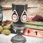 A pair of wooden teardrop-shaped earrings depicting a crystal ball and eye, hanging from a dark metal chalice in a dark academia setting.