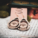 Handcrafted crystal ball earrings displayed on a "The Griffin and Stag" backing card atop pages of hand written calligraphy in a dark academia setting.