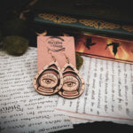 Handcrafted crystal ball earrings displayed on a "The Griffin and Stag" backing card atop pages of hand written calligraphy in a dark academia setting.