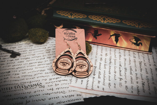 Handcrafted crystal ball earrings displayed on a "The Griffin and Stag" backing card atop pages of hand written calligraphy in a dark academia setting.