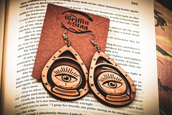 Handcrafted crystal ball earrings displayed on a "The Griffin and Stag" backing card atop the page of a fantasy hardcover book.