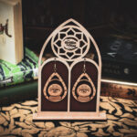Laser cut and engraved earrings featuring an eye with beams of light coming from it inside crystal ball within a teardrop frame hanging from a display stand in the shape of a gothic window in front of stacks of books.