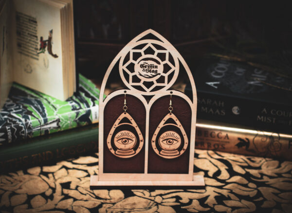 Laser cut and engraved earrings featuring an eye with beams of light coming from it inside crystal ball within a teardrop frame hanging from a display stand in the shape of a gothic window in front of stacks of books.