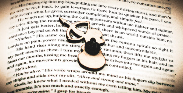 The back of a laser engraved raw birch plywood pin featuring a half-man, half-snail shooting a bow and arrow lying on top of the page of a book.