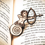 The front of a laser engraved raw birch plywood pin featuring a half-man, half-snail shooting a bow and arrow lying on top of the page of a book.