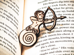 The front of a laser engraved raw birch plywood pin featuring a half-man, half-snail shooting a bow and arrow lying on top of the page of a book.