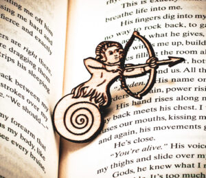 The front of a laser engraved raw birch plywood pin featuring a half-man, half-snail shooting a bow and arrow lying on top of the page of a book.