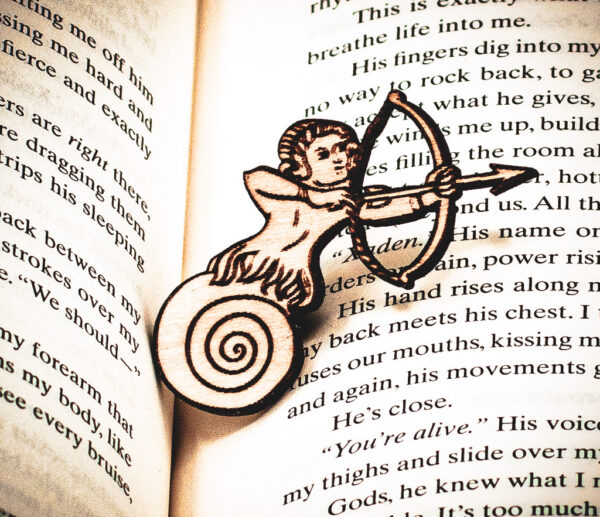 The front of a laser engraved raw birch plywood pin featuring a half-man, half-snail shooting a bow and arrow lying on top of the page of a book.