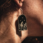 A close-up side profile of a person wearing a Star of Velaris earring, showcasing the silver-on-black mountain design against the sunlight.