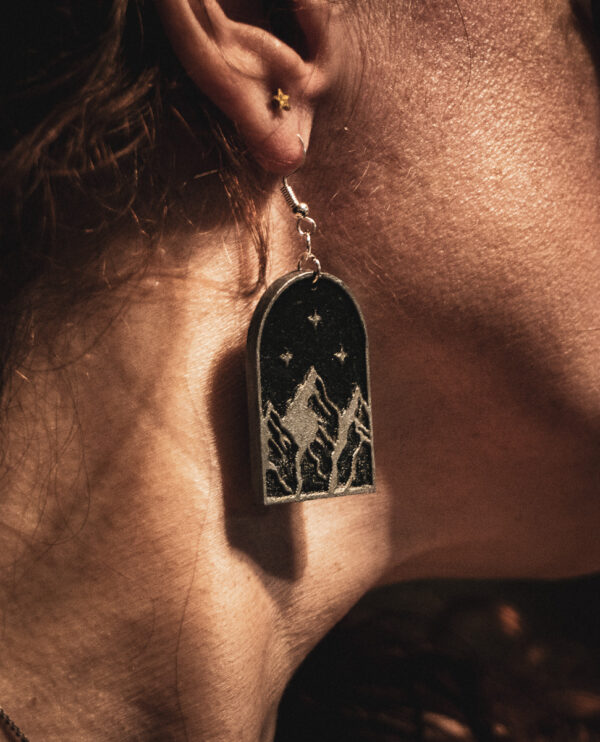 A close-up side profile of a person wearing a Star of Velaris earring, showcasing the silver-on-black mountain design against the sunlight.