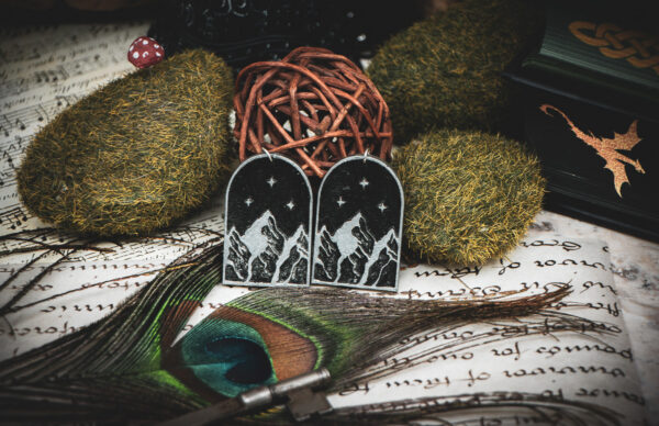 Handcrafted mountain earrings styled on a green book with gold Celtic knots, surrounded by mossy stones, a peacock feather, and a vintage key.