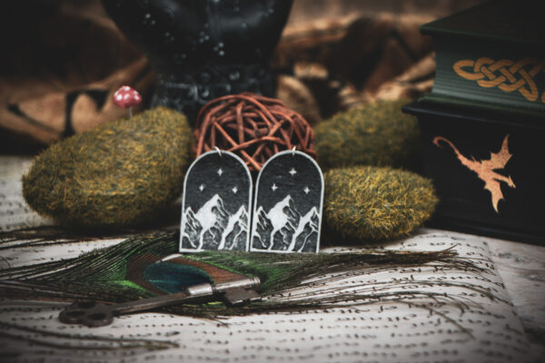 Handcrafted mountain earrings styled on a green book with gold Celtic knots, surrounded by mossy stones, a peacock feather, and a vintage key.