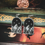 Laser cut and engraved earrings featuring an arch with the symbol of the Night Court, three mountain peaks with three stars framing the topmost peak, painted black and silver hanging between the pages of books.