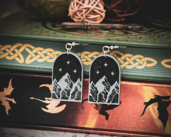 Laser cut and engraved earrings featuring an arch with the symbol of the Night Court, three mountain peaks with three stars framing the topmost peak, painted black and silver hanging between the pages of books.