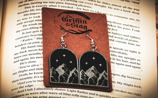 A pair of arch-shaped wooden earrings featuring an engraved silver mountain range under three stars, displayed on a brown "The Griffin and Stag" backing card over an open book.