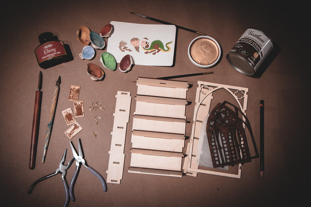 Paints, ink, stain, and tools laid out with partially assembled products on a kraft paper surface