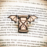 A pin featuring a winged hourglass engraved and cut from 1/8" raw birch plywood on top of the page of a book.