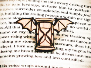 A pin featuring a winged hourglass engraved and cut from 1/8" raw birch plywood on top of the page of a book.