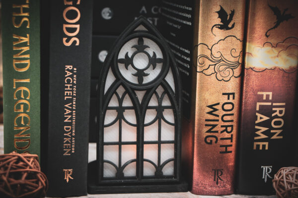 An architectural book nook lantern featuring laser-cut Gothic cathedral window tracery made from 1/8 inch birch plywood painted matte black between hardback fantasy books.